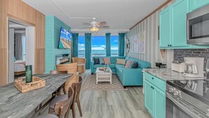 Een moderne keuken met blauwgroene kastjes, een eethoek met een houten tafel en stoelen, een woonkamer met een blauwgroene bank en een televisie, en uitzicht op de oceaan.