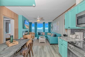 A modern kitchen with teal cabinets, a dining area with a wooden table and chairs, a living room with a teal sofa and a TV, and a view of the ocean. - Yachtsman S606 2 BR (Myrtle Beach)