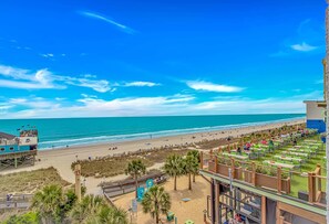Vista de la playa con un paseo marítimo, palmeras y una torre de salvavidas, y un edificio con terraza.