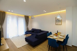 Ein modernes Wohnzimmer mit einem dunkelblauen Ecksofa, einem Essbereich mit blauen Stühlen und einem Tisch für vier Personen sowie einem großen Fenster mit transparenten Vorhängen.