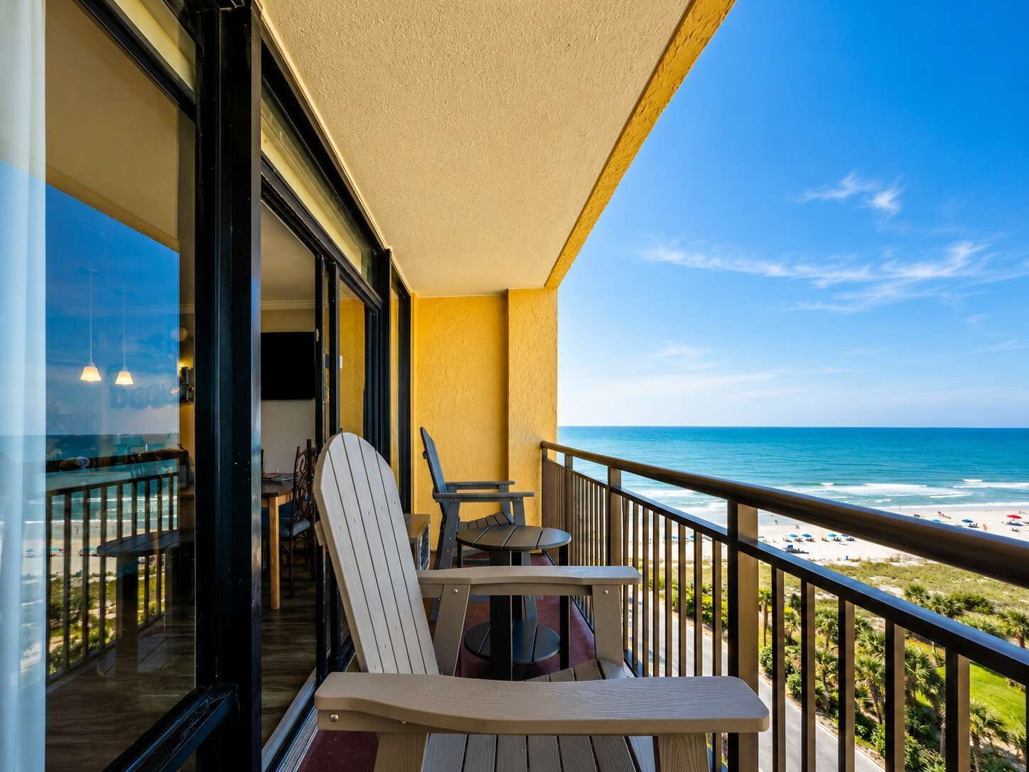 A balcony with a view of the beach, a table, two chairs, and a glass door leading to an interior space.