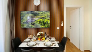 Un coin repas avec une table dressée pour quatre personnes, une horloge et un grand écran de télévision diffusant un paysage pittoresque.