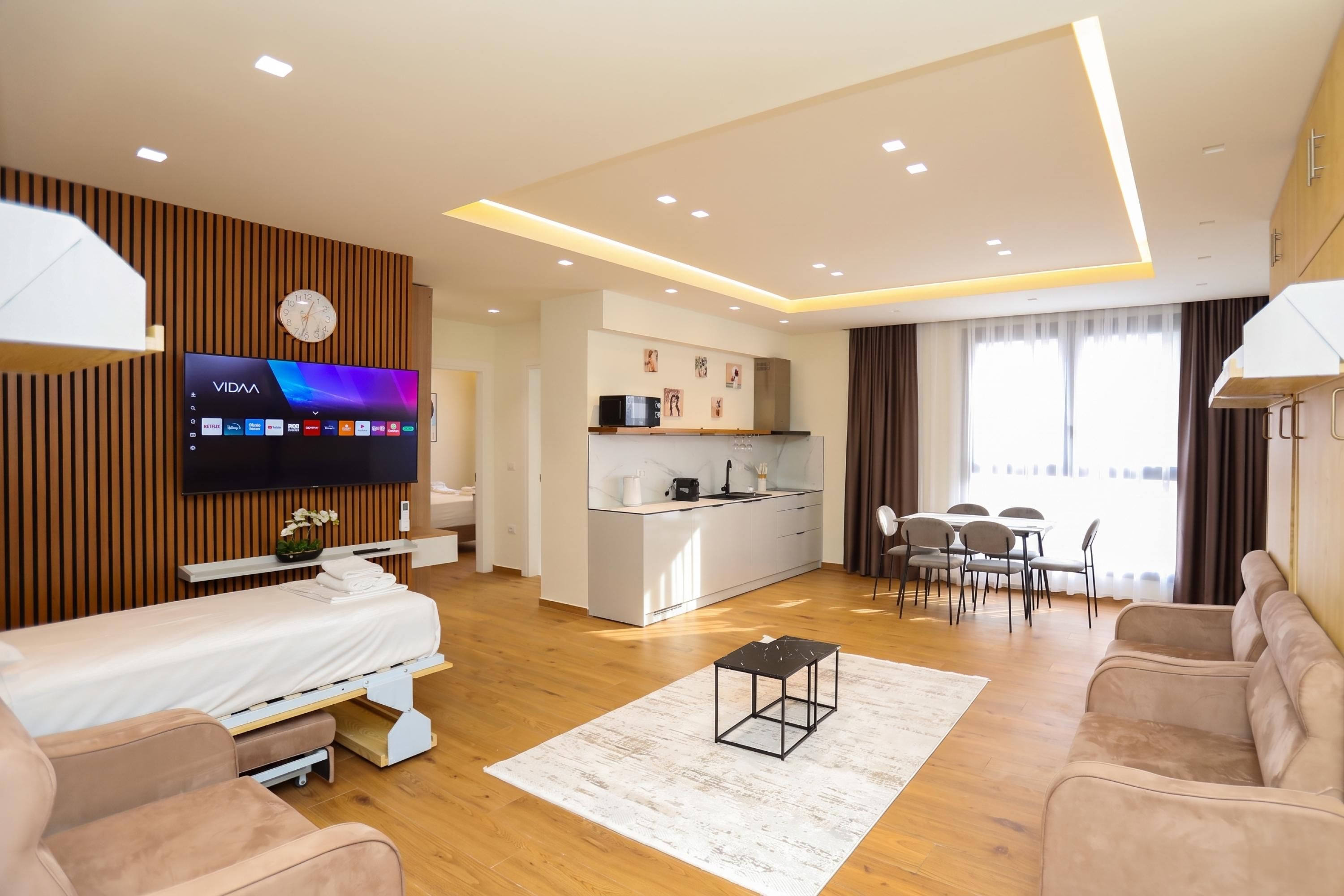A modern hotel room with a flat-screen TV, a bed, a sofa, a dining table, and a kitchen area.