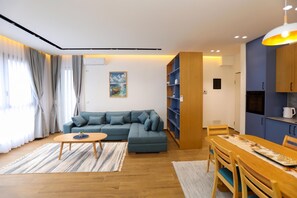 A modern living room with a sectional sofa, a dining table with chairs, a kitchen area, and a large window with curtains. - La Dimora Azzurra, Vlore, Albania (Vlore)