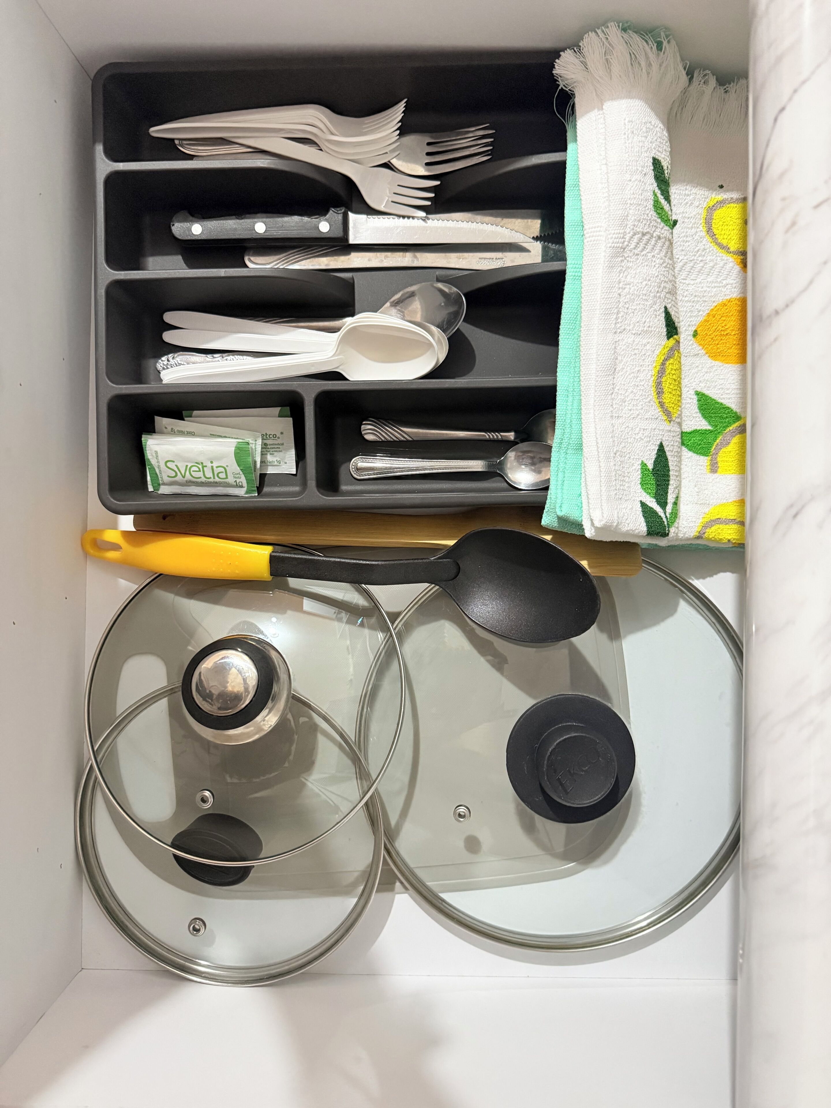 A kitchen drawer with utensils, a knife, a spoon, a spatula, a lemon-patterned towel, and a yellow-handled knife.