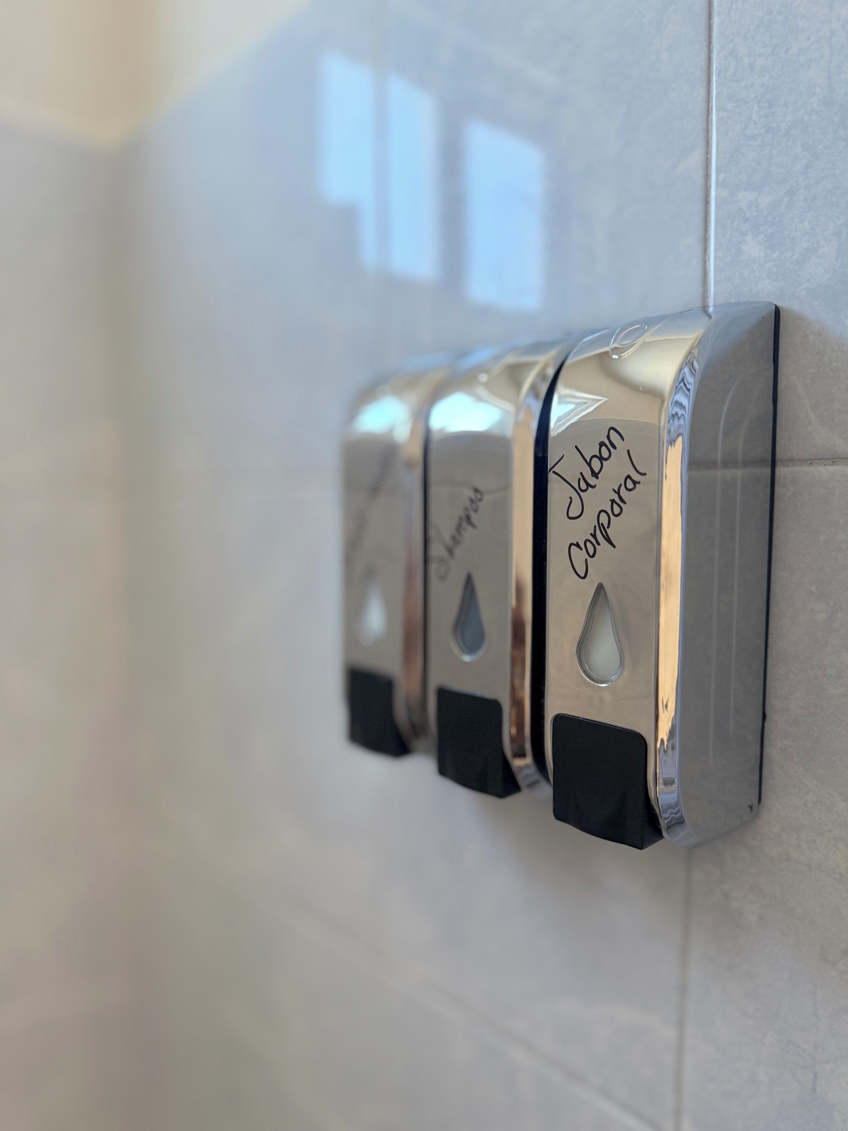 Eine Reihe metallischer Seifenspender mit der Aufschrift 'Jabon Corporal' an einer gefliesten Wand.