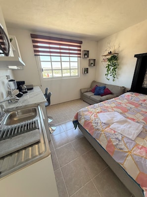 A compact room with a kitchenette, a bed with a patterned quilt, a window with blinds, and a hanging plant. - Cozy refuge in Puebla: comfort, style and ideal location, Pet friendly. (Heroica Puebla de Zaragoza)