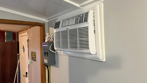 Een kamer met een witte airconditioningunit aan de muur, een deur met houten frame en een elektrisch paneel dat aan de wand is bevestigd.