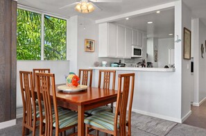 A dining area with a wooden table and chairs, a kitchen with white cabinets, and a window with a view of green foliage. - Beach Paradise Getaway at North Coast Village G18 (Oceanside)