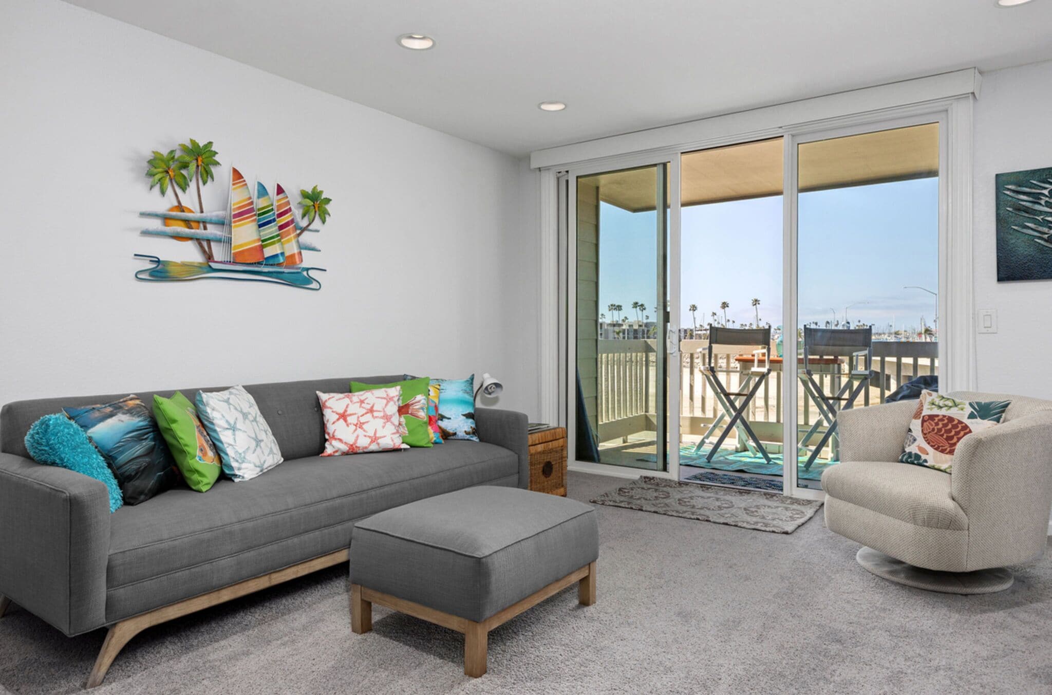 Un salon moderne avec un canapé gris, des coussins colorés, un pouf gris, un fauteuil beige et une grande porte-fenêtre coulissante donnant sur un balcon avec vue sur des palmiers et le paysage urbain.