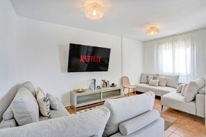 Una sala de estar moderna con un televisor de pantalla plana montado en la pared, un conjunto de sofás, una mesita auxiliar y una ventana con cortinas.