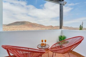 Een balkon met twee rode rieten stoelen, een klein rond tafeltje met een bord eten en twee drankjes, en uitzicht op een woestijnlandschap met bergen.