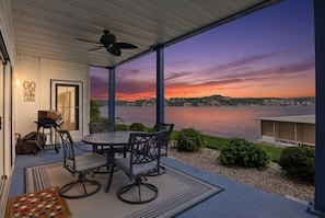 A covered patio with a table and chairs, a ceiling fan, and a view of a lake at sunset. - Lakefront Updated Condo at 2mm w/ 2 Pools, Hot Tub, Boat Slip (Lake Ozark)