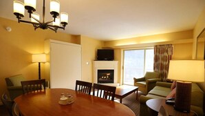 Habitación de hotel con una mesa de comedor, sillas, un sofá, un televisor, una chimenea y una ventana con cortinas.