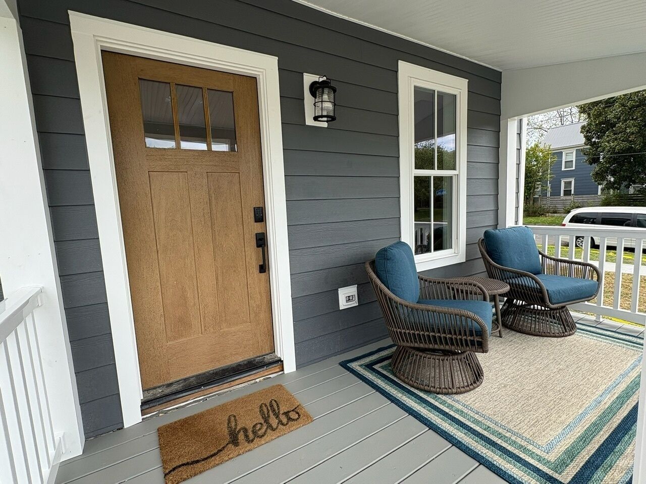 A porch with a wooden door, two wicker chairs, a small table, and a 'hello' mat.