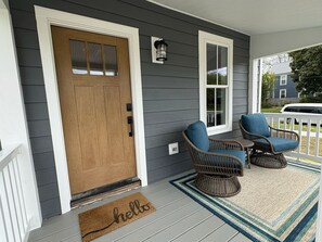 Eine Veranda mit einer Holztür, zwei Korbstühlen, einem kleinen Tisch und einer Fußmatte mit dem Schriftzug 'hello'.