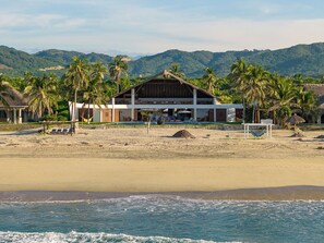 A beachfront hotel with a thatched-roof structure, a pool, and palm trees.