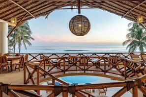 A beachside lounge with a pool, wooden furniture, and a thatched roof.