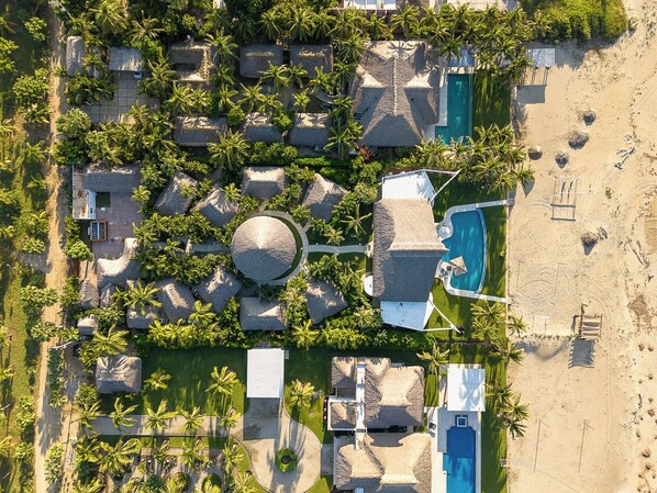 An aerial view of a resort with multiple thatched-roof structures, a central circular building, a swimming pool, and a beach area.