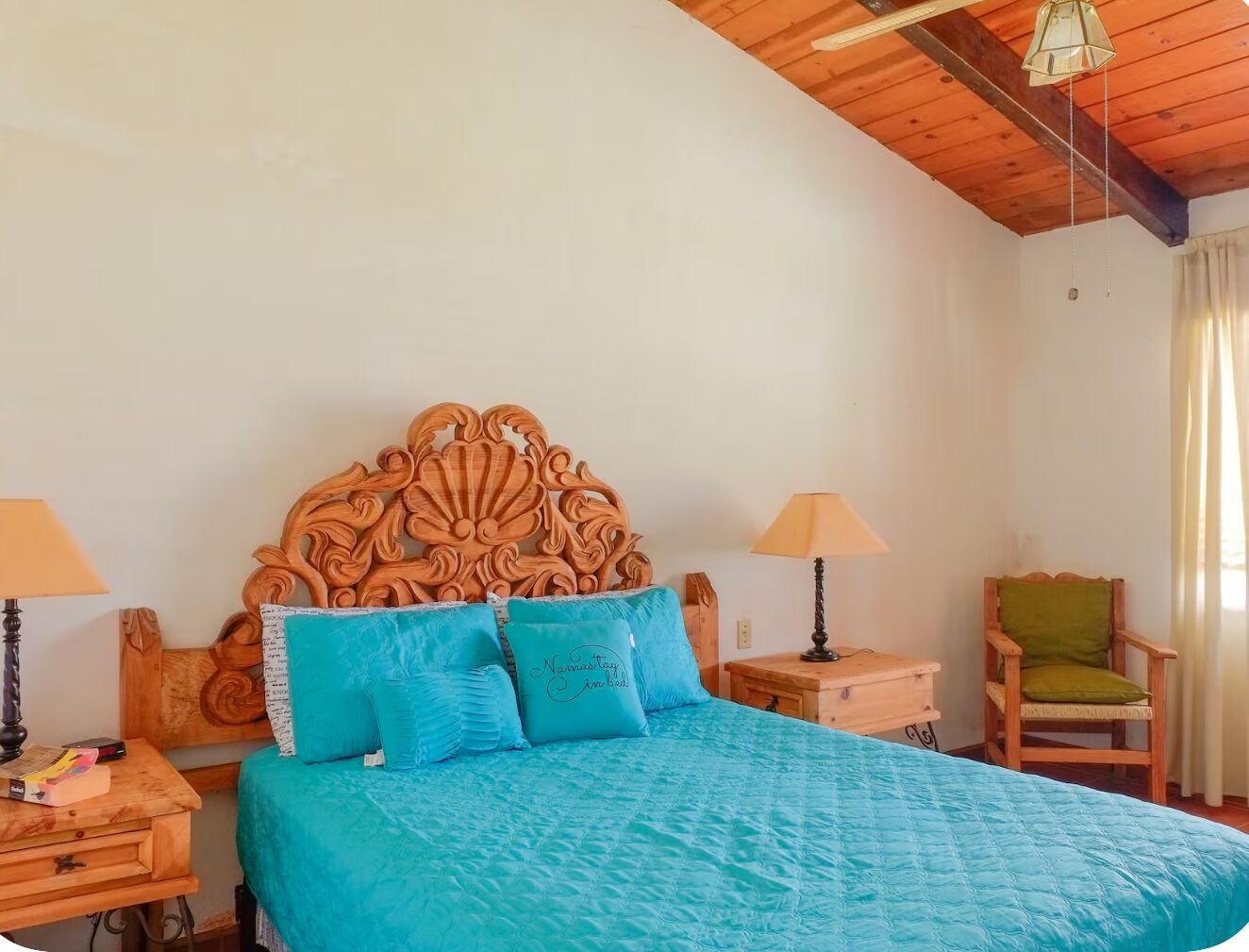 A bedroom with a wooden headboard, a bed with a blue quilt, two bedside tables with lamps, and a chair.