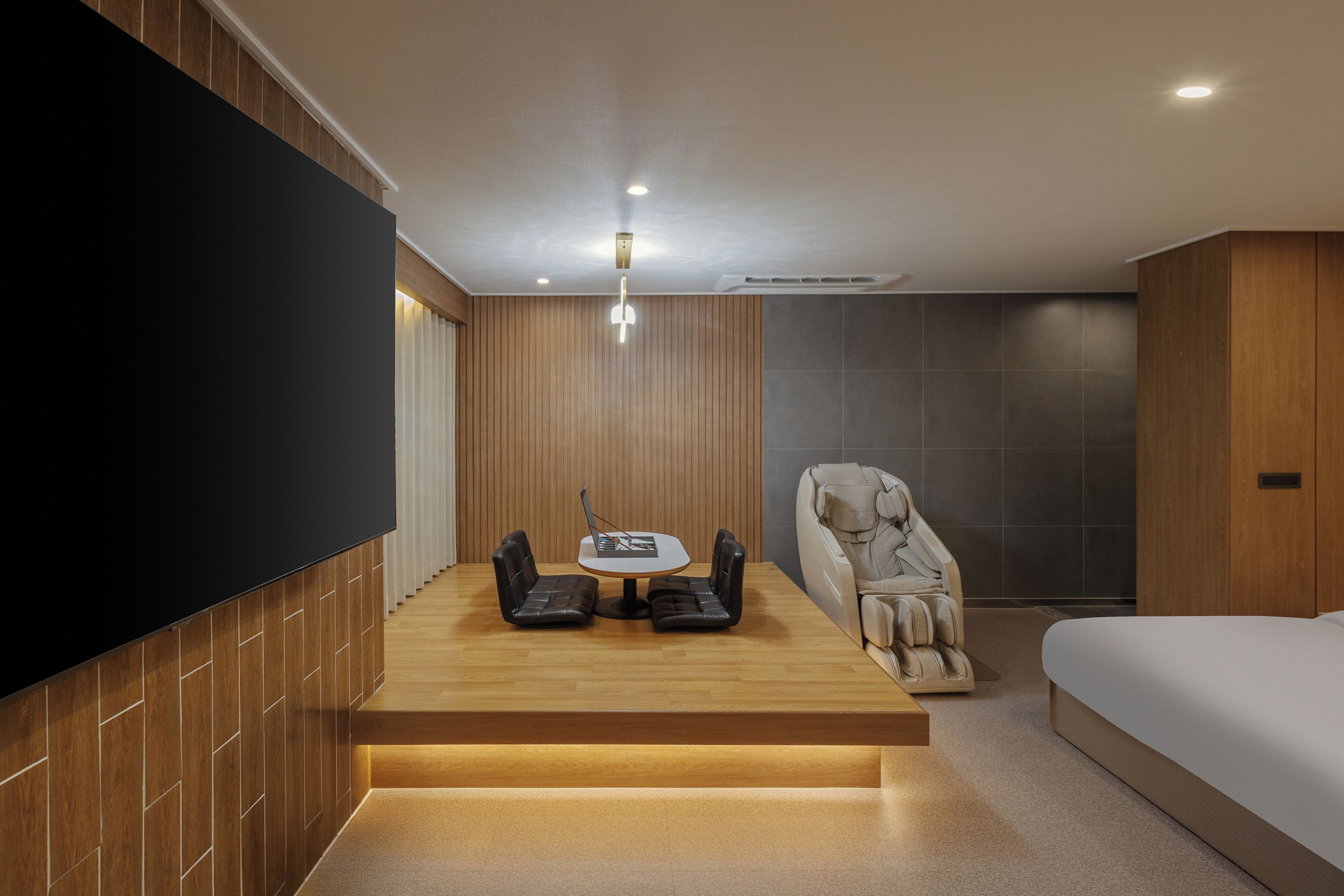 Ein modernes Zimmer mit Flachbildfernseher, Massagesessel, kleinem Tisch mit Laptop und Bett.
