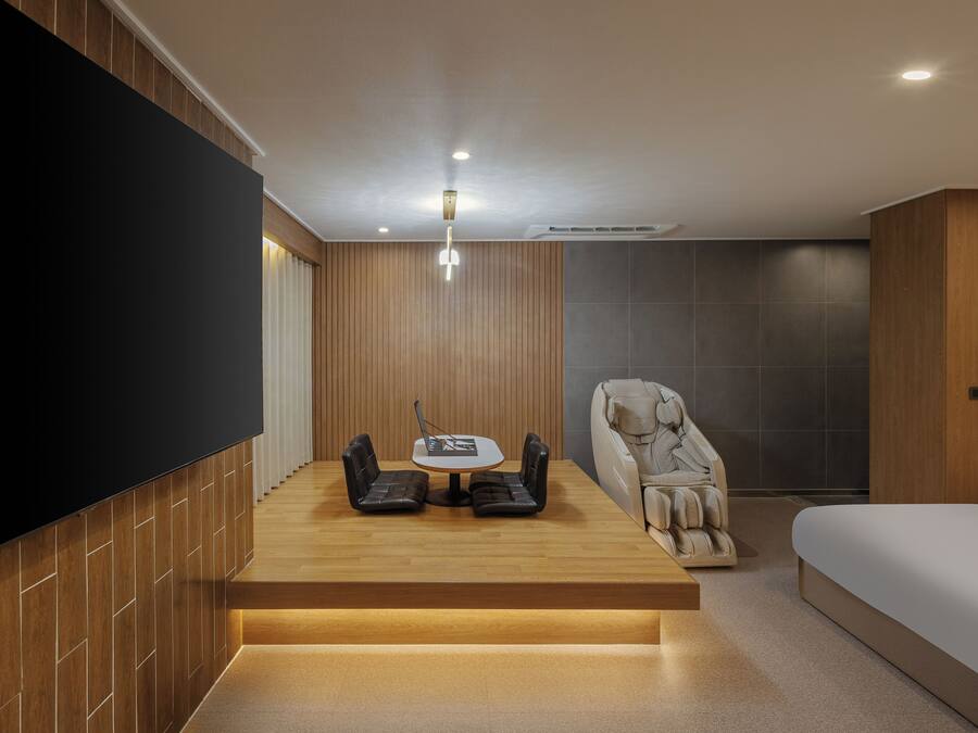 Ein modernes Zimmer mit Flachbildfernseher, Massagesessel, kleinem Tisch mit Laptop und Bett.