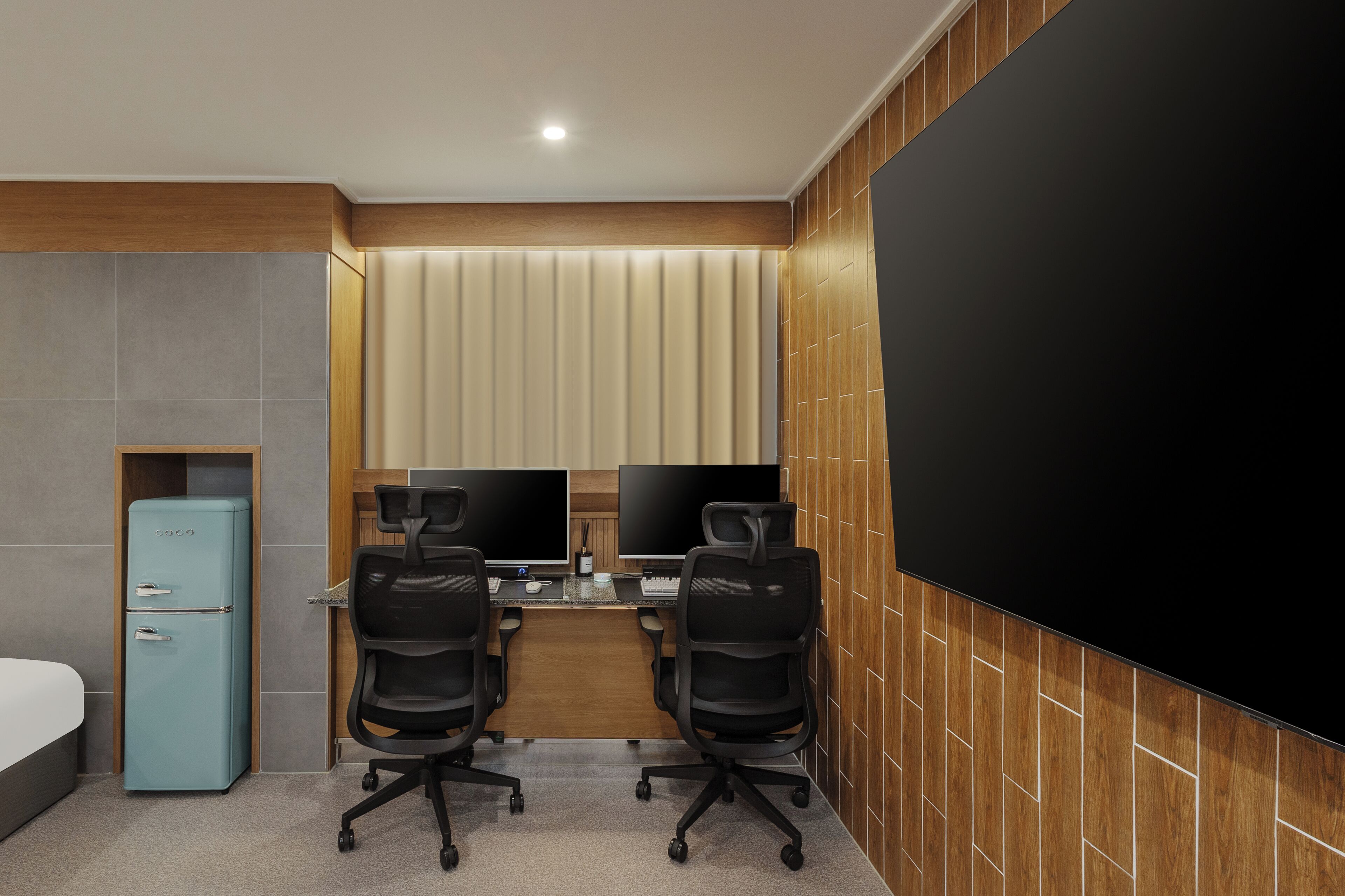 A modern hotel room with a large bed, a desk with two monitors, two office chairs, and a large TV screen.