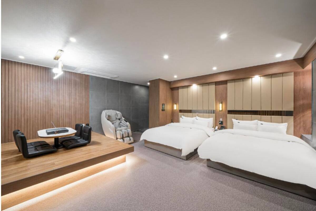 Ein modernes Hotelzimmer mit Schreibtisch aus Holz, zwei Betten, einem Massagesessel und einem großen Spiegel.