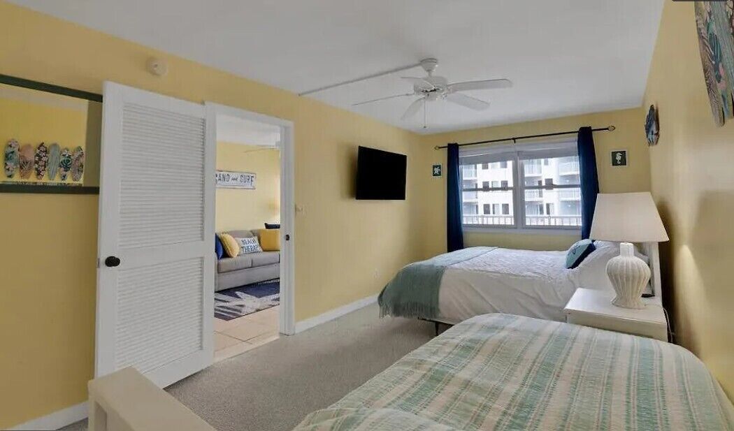 Ein Schlafzimmer mit zwei Betten, einem Deckenventilator, einem Fernseher und einem Fenster mit Vorhängen.