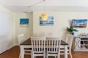 Un coin repas avec une table en bois et des chaises blanches, un luminaire suspendu et deux tableaux accrochés au mur.