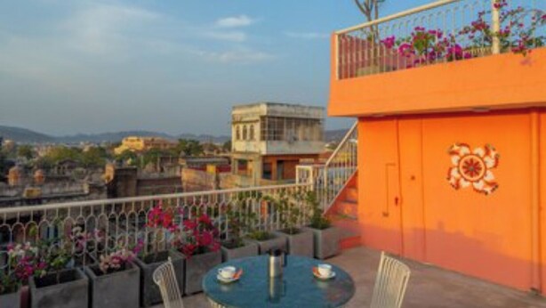 A rooftop terrace with a round table and chairs, a railing with flowers, and a view of buildings in the background. - Sheel Mahal Near Hawa Mahal (Jaipur)