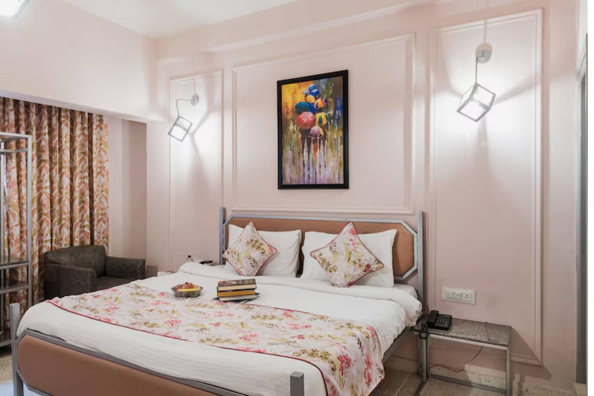 A bedroom with a bed, a chair, a painting, and a desk with a lamp.