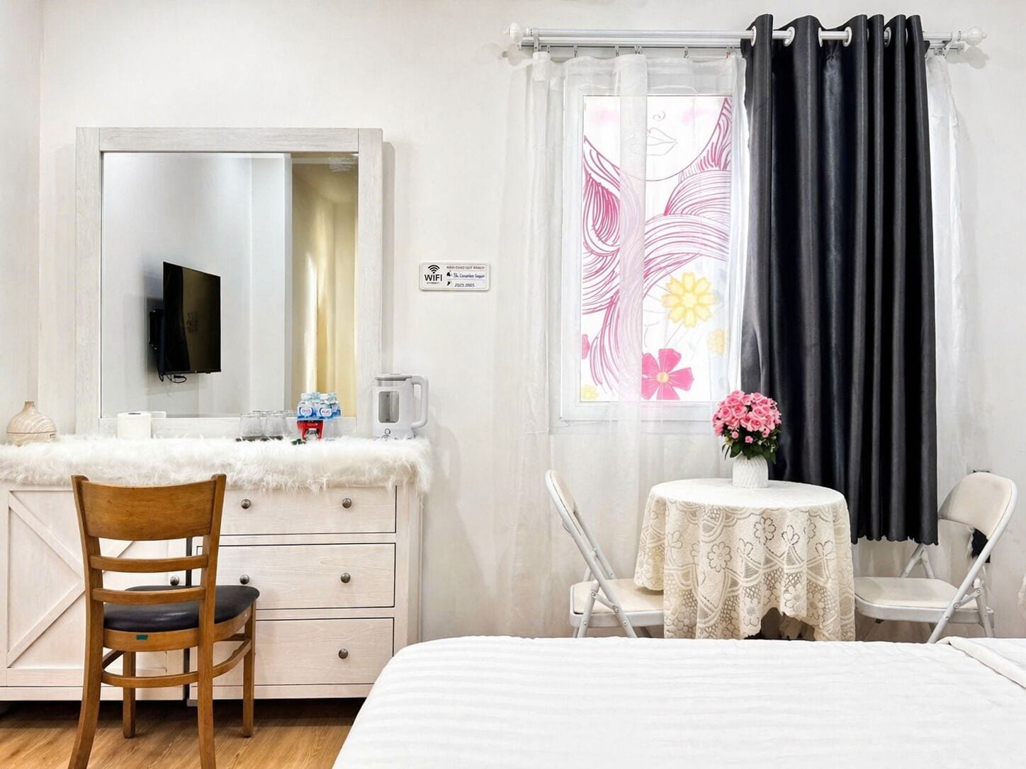 Ett hotellrum med en säng, en byrå med en spegel, en tv, ett litet bord med en vas med blommor och ett fönster med gardiner.