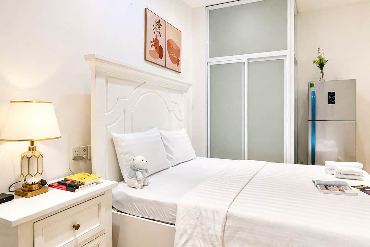 A hotel room with a bed, bedside table, lamp, and a refrigerator.