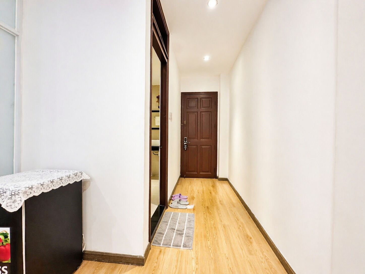 A hallway with a wooden floor, a dark door at the end, and a small table with a lace cover on the left.