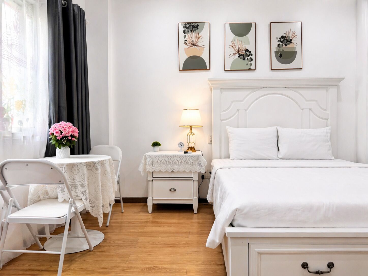 A white bedroom with a bed, a nightstand, a chair, a table, and three wall-mounted paintings.
