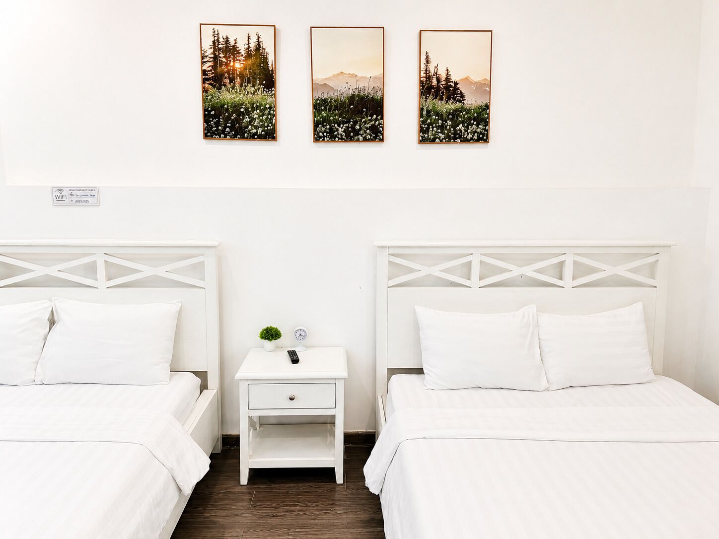 Habitación de hotel con dos camas, una mesita de noche con una planta y un reloj, y tres cuadros en la pared.