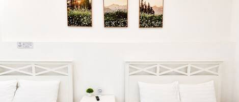 Habitación de hotel con dos camas, una mesita de noche con una planta y un reloj, y tres cuadros en la pared.