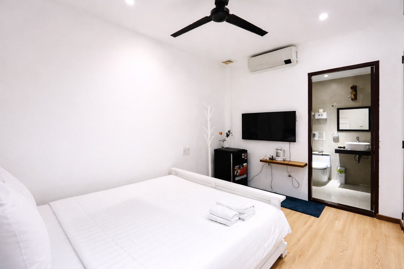 A modern hotel room with a bed, ceiling fan, air conditioning unit, TV, and a bathroom with a sink and mirror.