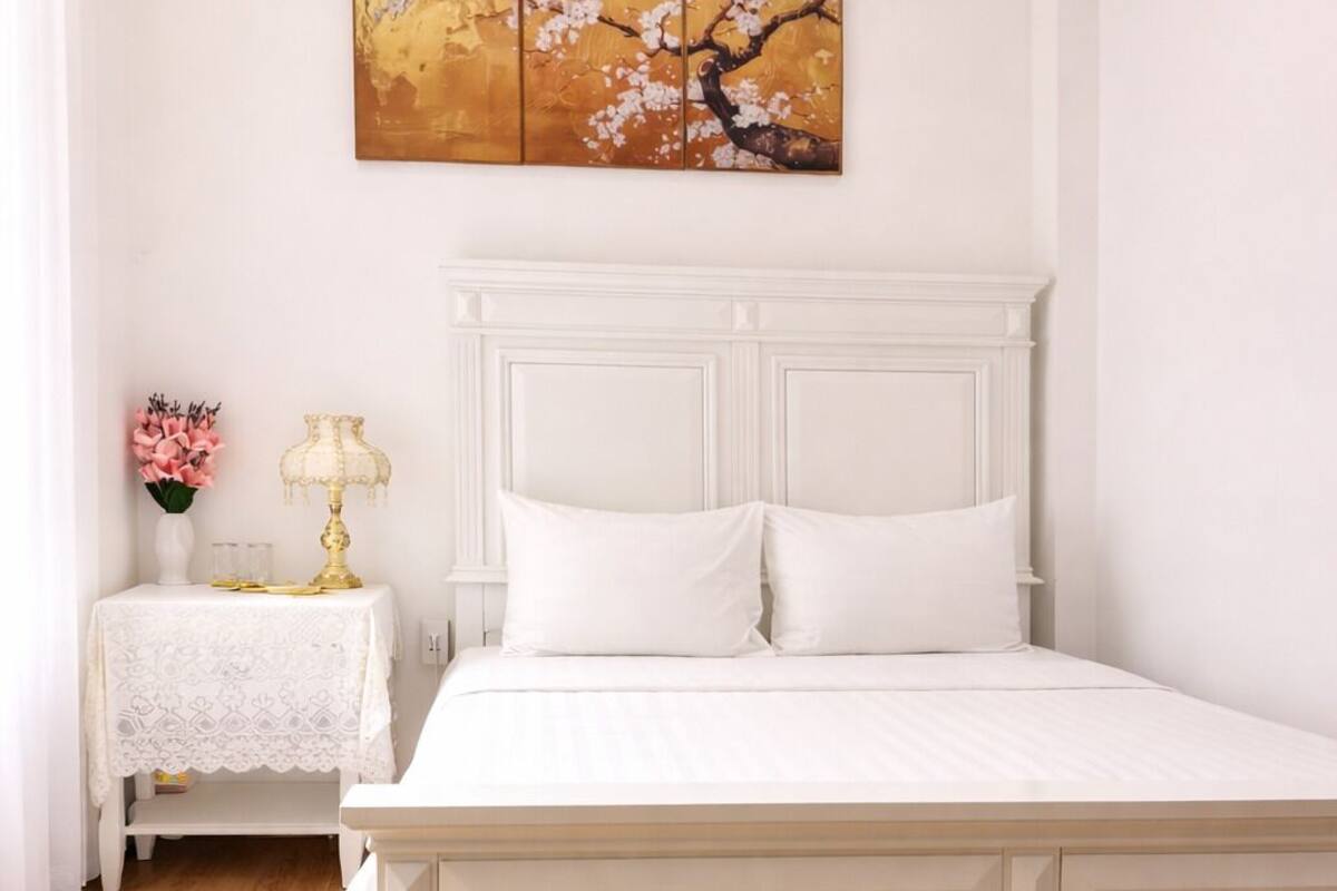 A white four-poster bed with a white headboard, a bedside table with a lamp, and a wall-mounted painting.