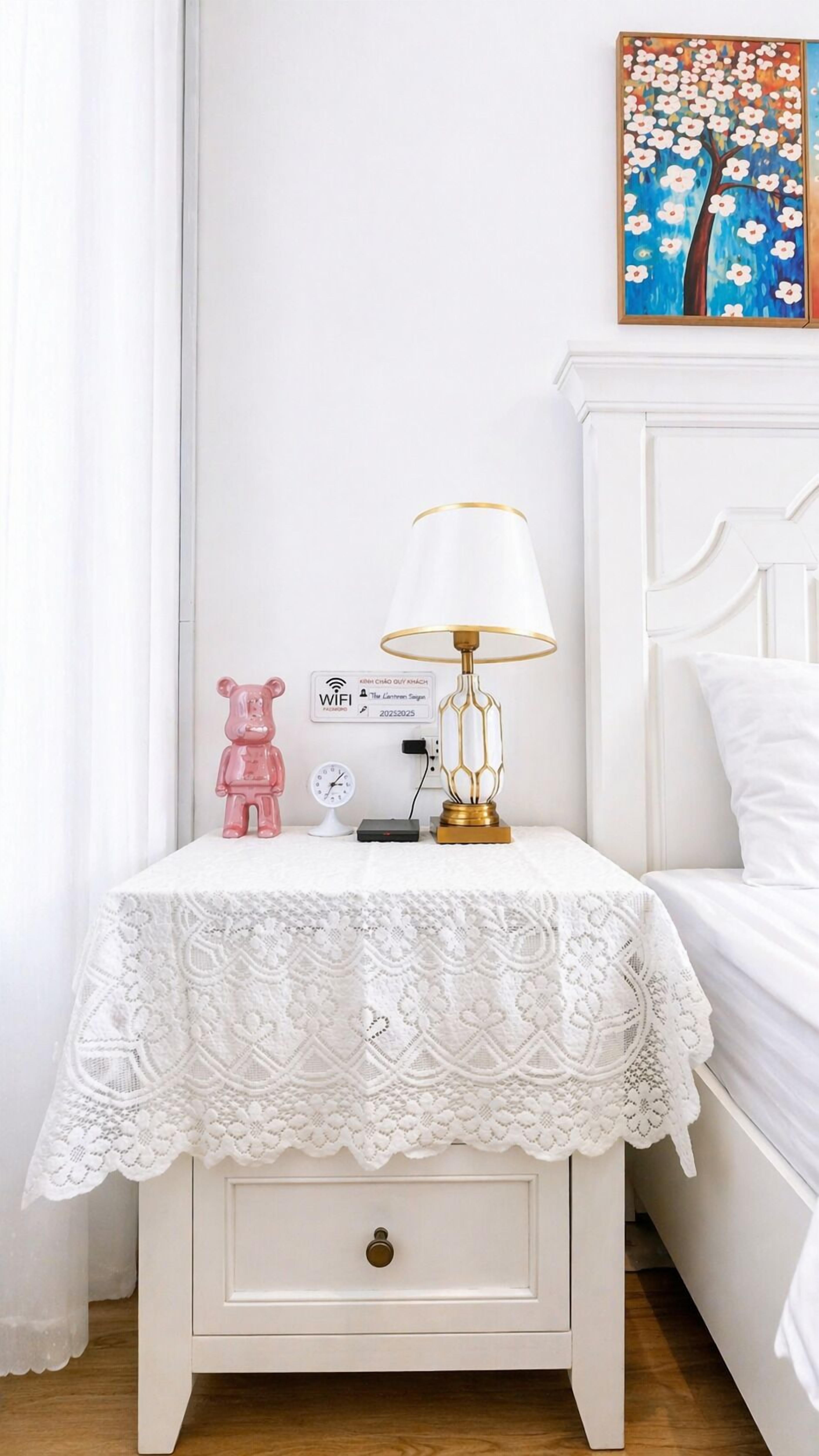 Una mesita de noche blanca con mantel de encaje, una lámpara dorada, una figurita de un oso rosa, un cartel de WiFi y un reloj.