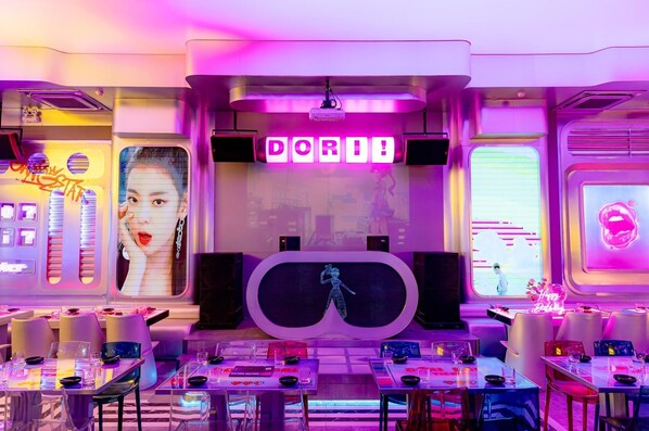A futuristic interior with neon signage, a central projection screen, and tables set for dining. - Movida Stay - Bui Vien Walking Street, District 1 (Thanh Pho Ho Chi Minh)