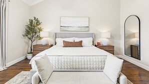 A bedroom with a large bed, bedside lamps, a nightstand, a mirror, and a potted plant.