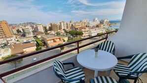 Un balcón con una mesa blanca y sillas a rayas, con vistas a un paisaje urbano que incluye edificios y una masa de agua.