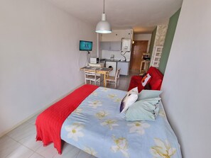 Ein Schlafzimmer mit einem Bett, einer roten Decke, einem Schreibtisch mit Stuhl, einem Fernseher und einer kleinen Küchenzeile im Hintergrund.