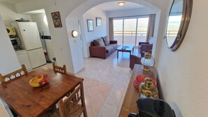 Un espacio de estar con una mesa de comedor, sillas, un sofá y una mesa auxiliar. Hay una cocina con refrigerador, microondas y estufa. La habitación tiene un ventanal con vistas a la playa.