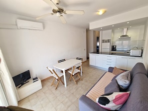 A living room with a sofa, a dining table with chairs, a ceiling fan, a TV, and a kitchen area with a refrigerator and microwave. - APARTMENT BADIA 24 (Empuriabrava)