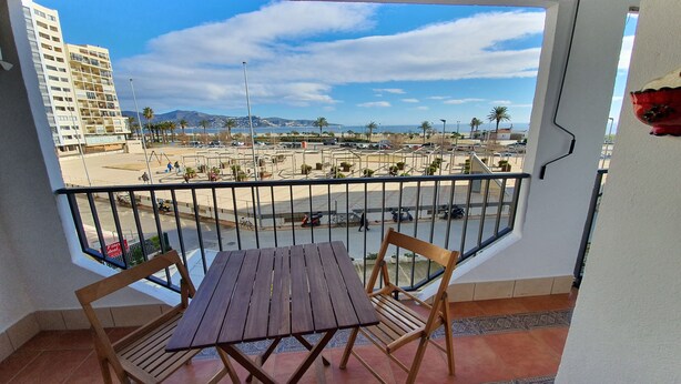 Un balcon avec une table et des chaises en bois, donnant sur une plage et des immeubles.