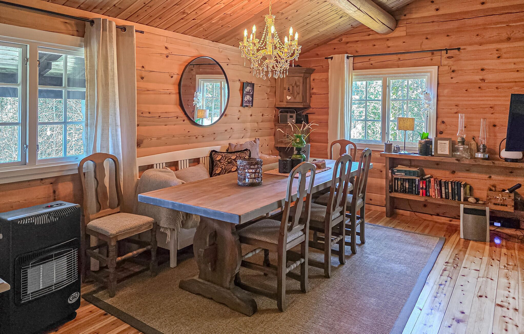 A cozy cabin interior with a wooden dining table, chairs, a chandelier, a fireplace, and a bookshelf.