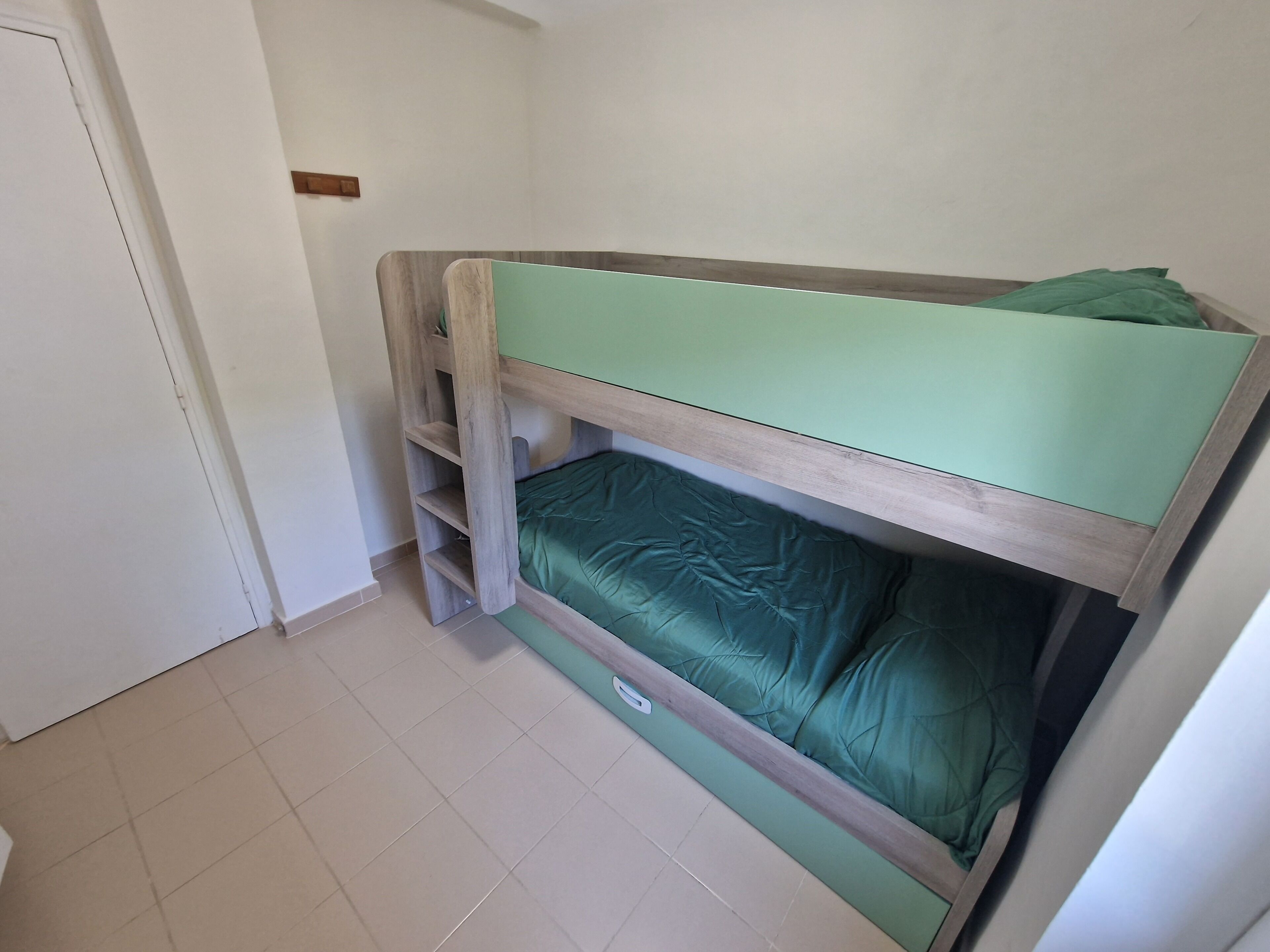 Una cama litera con colchón verde y estructura de madera en una habitación con paredes blancas y suelo de baldosas.